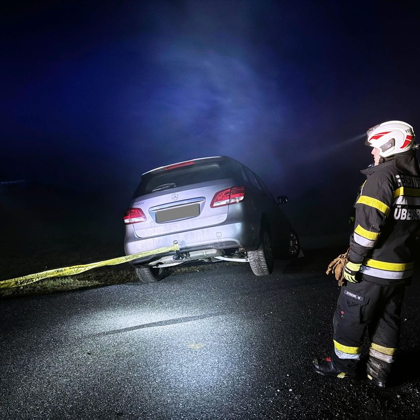 Ein Feuerwehrmann steht neben einem geparkten Auto und benutzt eine Taschenlampe. Das Auto steht am Straßenrand mit gelbem Band. Die Szene ist nachts.