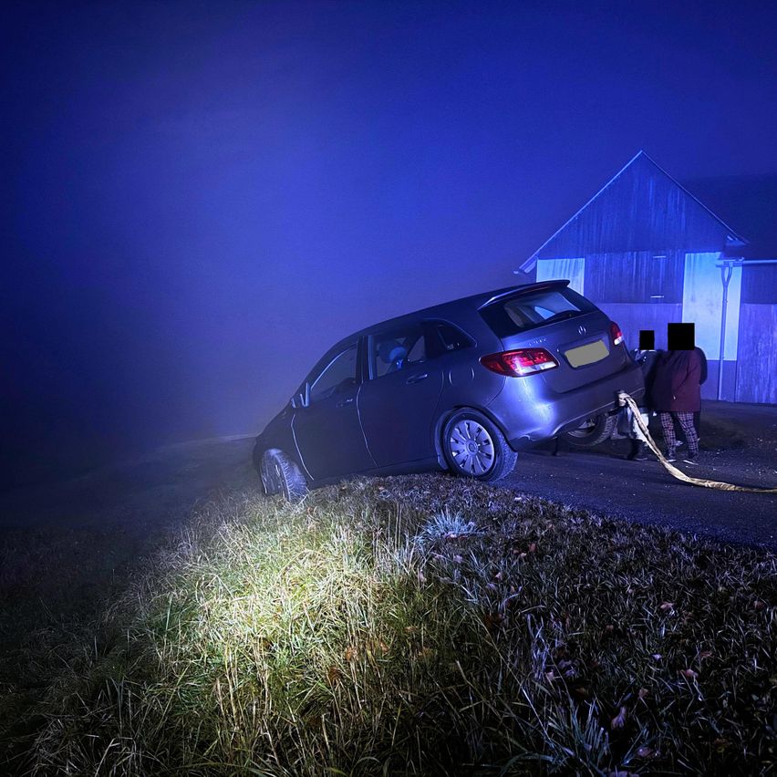 Ein Auto steht nachts am Straßenrand, wobei zwei Personen es ziehen. Das Auto ist grau und hat ein gelbes Nummernschild. Die Gegend ist neblig.