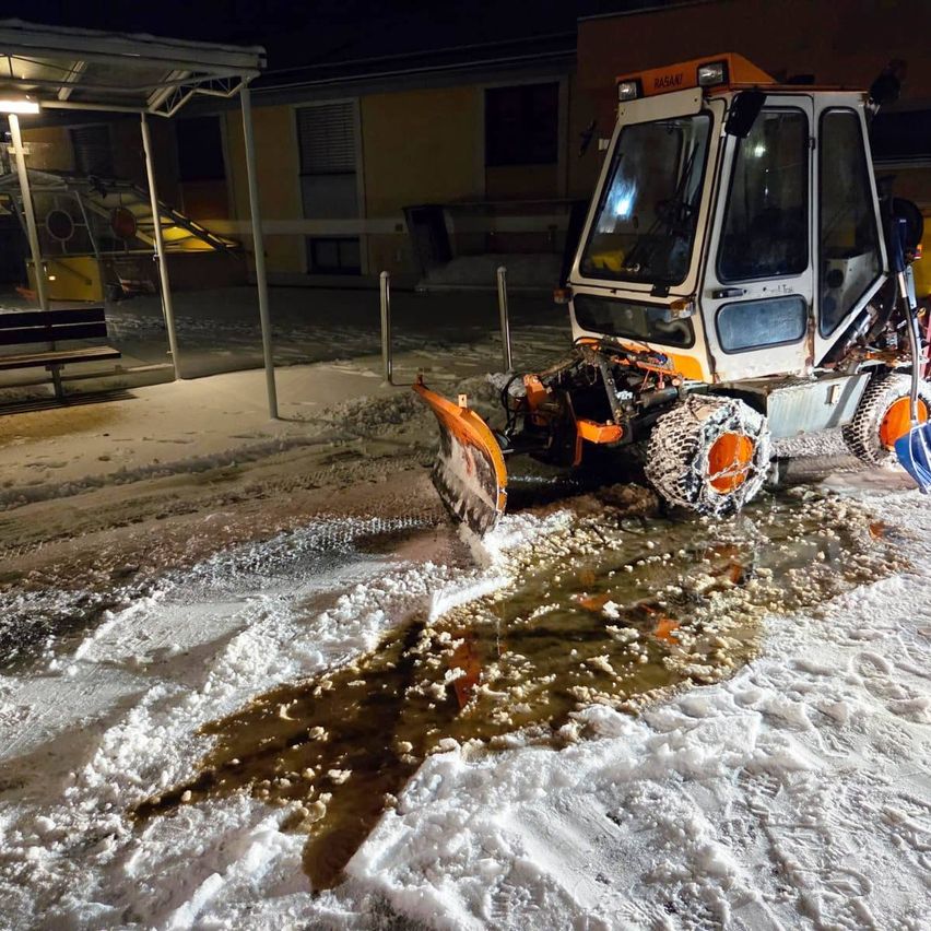 Ein Schneepflug räumt bei Nacht Schnee vor einem Gebäude. Der Schnee ist dick und nass, mit einer sich bildenden Pfütze.