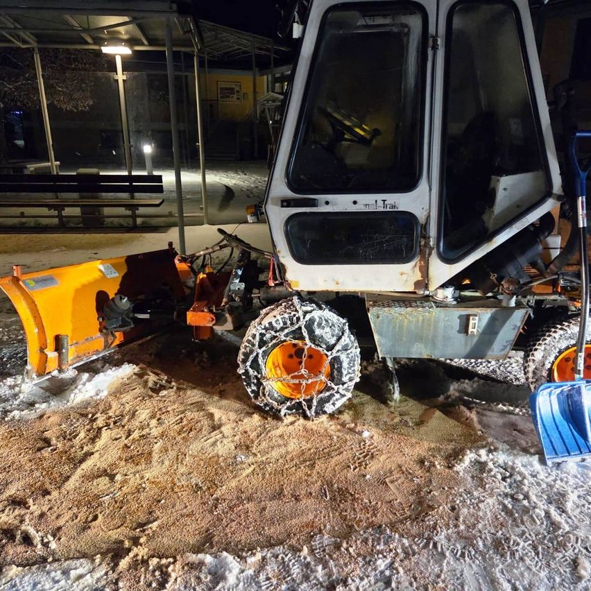 Ein Schneepflug mit gelber Frontschaufel steht im Schnee neben einer blauen Schneeschaufel. Dahinter befindet sich ein Gebäude mit einer Bank und einer Laterne.