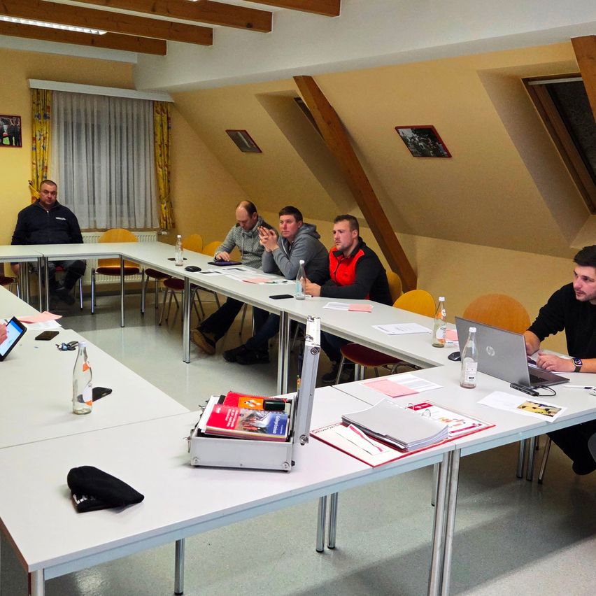 Fünf Männer sitzen um einen Konferenztisch herum mit Laptops und Büchern. Ein Mann gestikuliert und ein anderer schaut auf seinen Laptop.