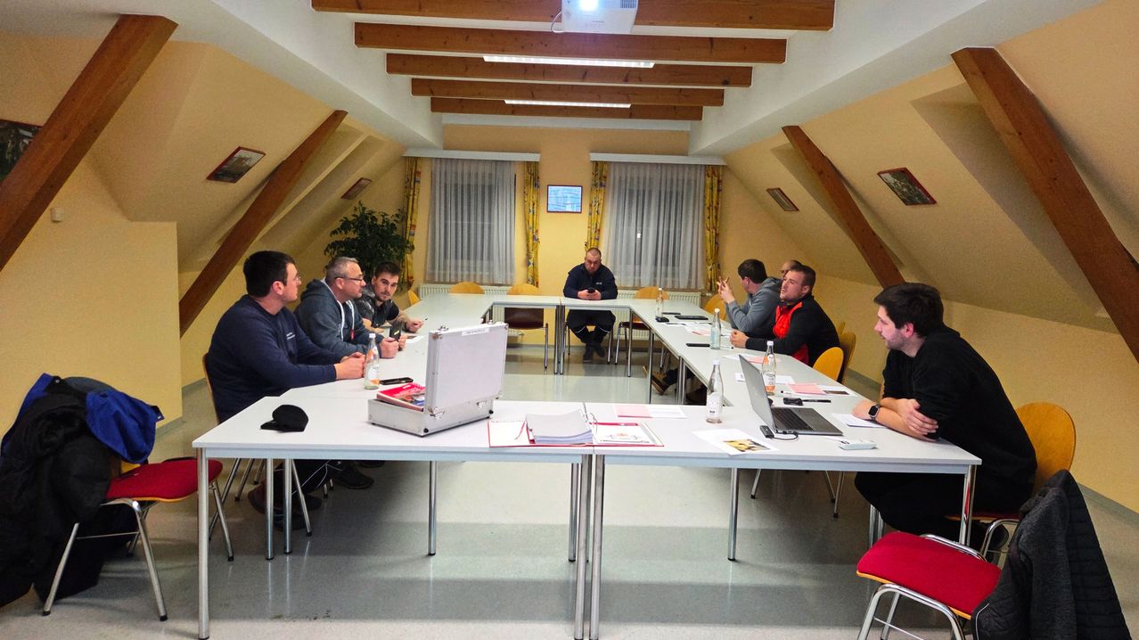 Eine Gruppe von Menschen sitzt in einem Konferenzraum um einen Tisch herum, mit Laptops und Papieren. Ein Mann steht vorne und spricht zu ihnen. Holzbalken säumen die Decke, und Vorhänge schmücken die Fenster.