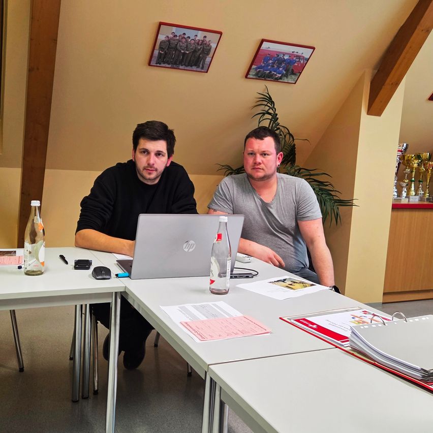 Zwei Männer sitzen an einem Tisch in einem Raum mit einem Laptop und einer Flasche. Einer trägt ein schwarzes Shirt, der andere ein graues T-Shirt. Papiere, eine Maus und Trophäen sind in der Nähe.