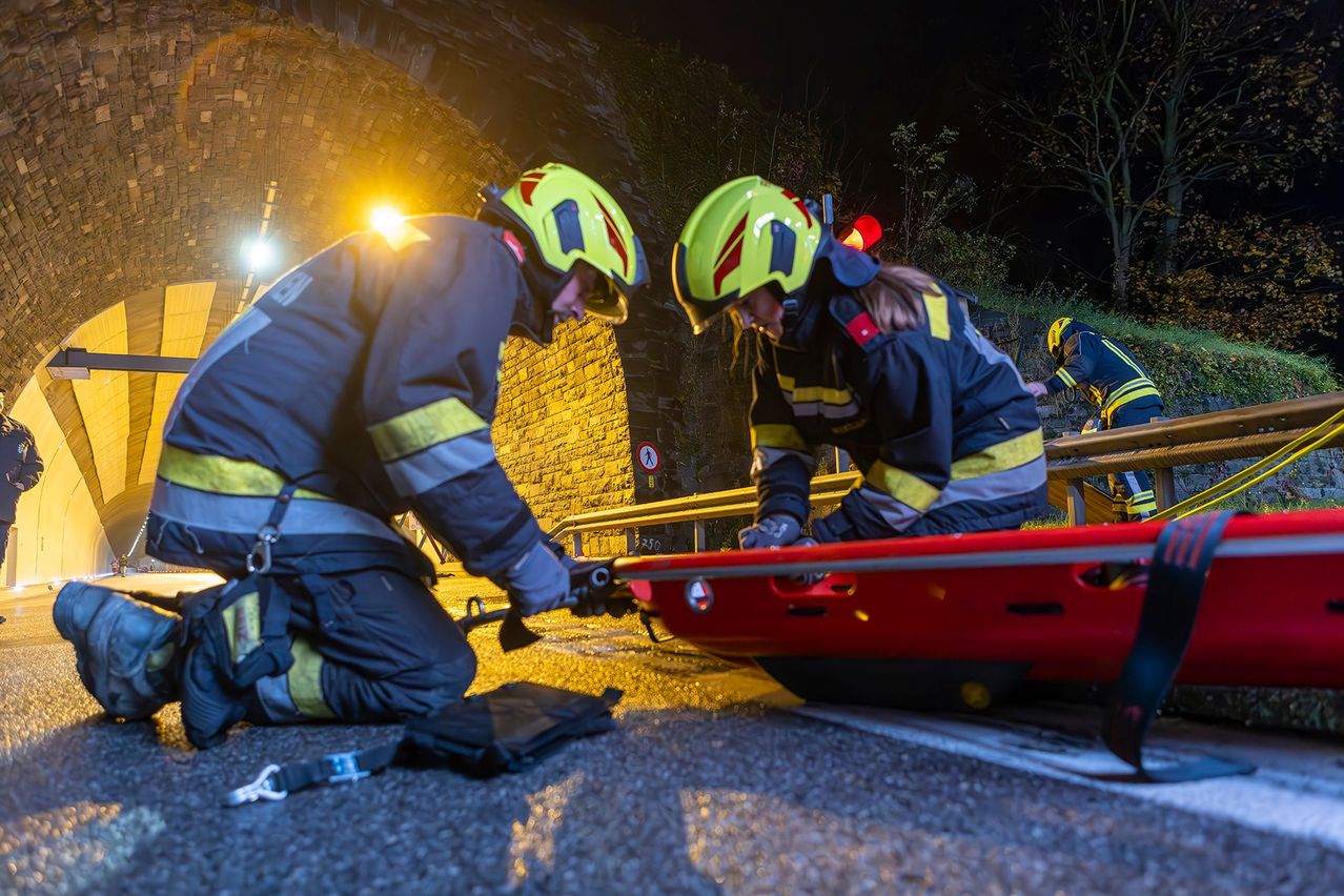 Zwei Feuerwehrleute in Helmen sichern eine rote Trage auf einer Straße, ein weiterer Feuerwehrmann steht dahinter. Die Szene spielt sich nachts unter einem Tunnel ab.