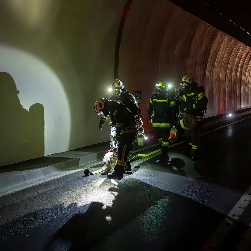 Feuerwehrleute in Helmen befinden sich in einem Tunnel mit hellen Lichtern, einer beugt sich herunter, um jemandem am Boden zu helfen.