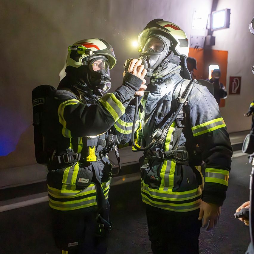 Zwei Feuerwehrleute in Schutzkleidung, einer justiert die Maske des anderen, in einem Tunnel mit orangefarbener Wand und Ausfahrtsschild.
