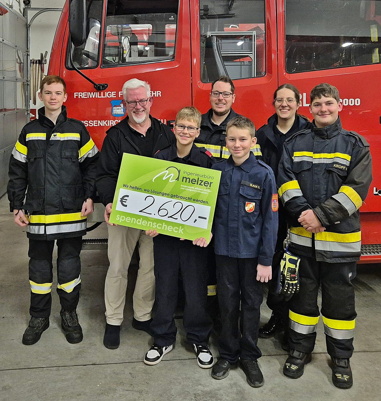 Eine Gruppe von Menschen, darunter zwei Kinder, steht vor einem roten Feuerwehrauto. Ein Mann hält einen grünen Scheck über 2.620 Euro.