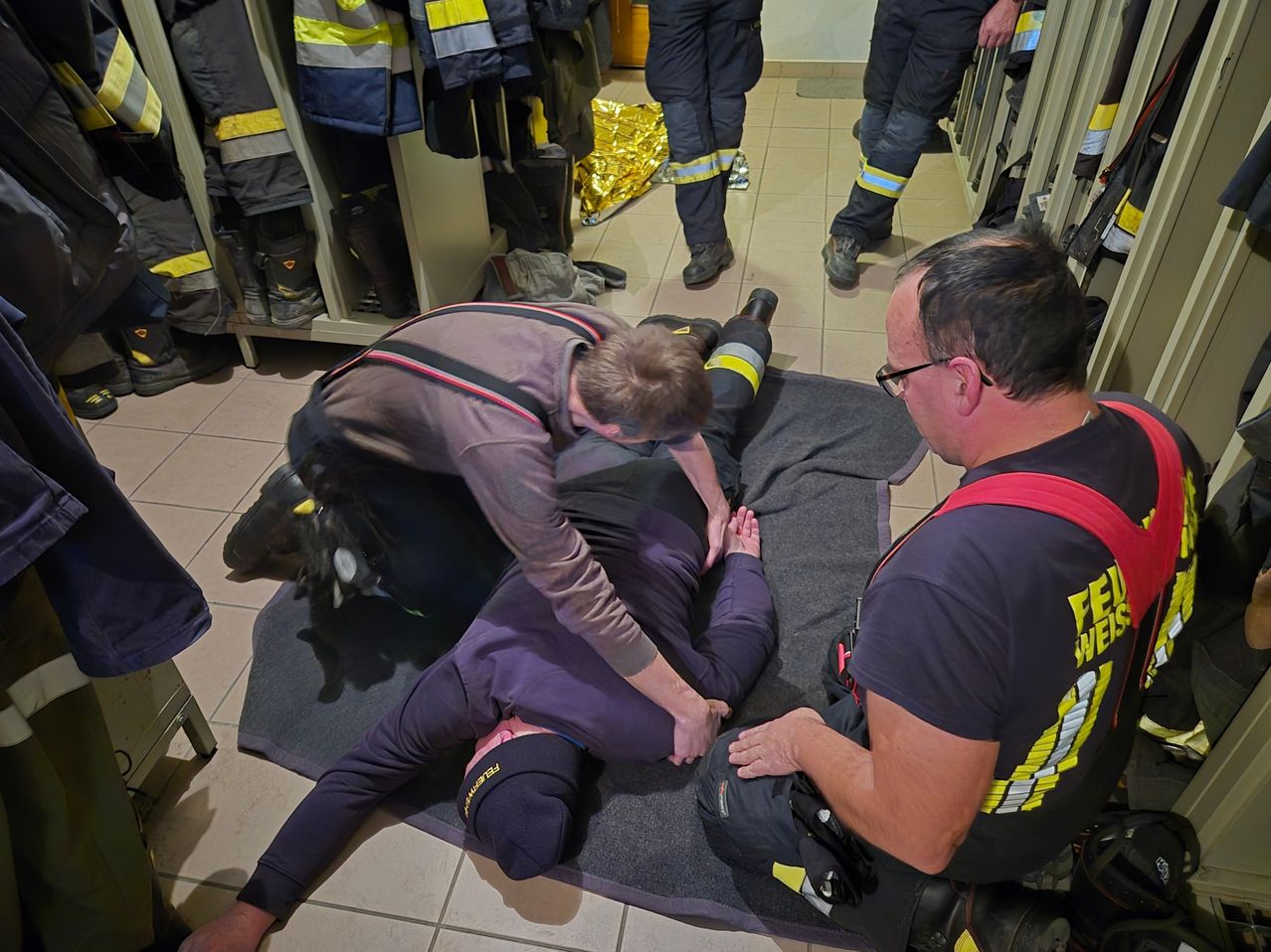 Eine Person, die auf einer Matte liegt, erhält von zwei Feuerwehrleuten medizinische Hilfe, von denen einer ein medizinisches Gerät hält. Sie befinden sich in einem Umkleideraum, in dem andere Feuerwehrleute stehen.