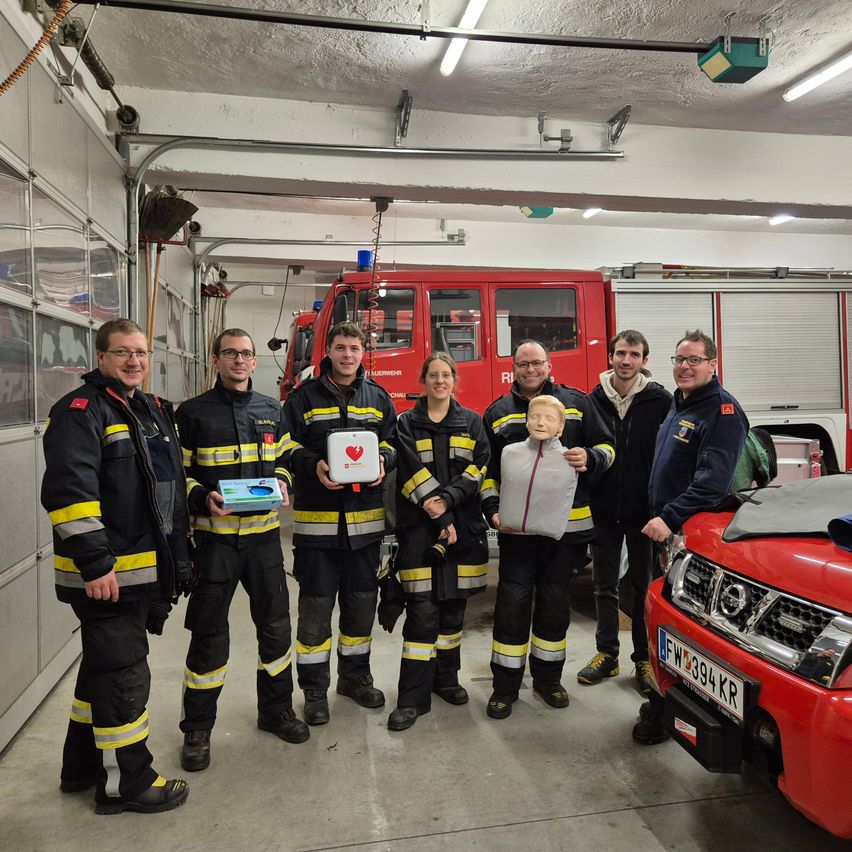 Eine Gruppe von Feuerwehrleuten in Uniformen steht in einer Garage und lächelt für ein Foto mit einem Feuerwehrwagen im Hintergrund. Ein Mann hält eine Box und ein anderer ein Puppenmannequin.