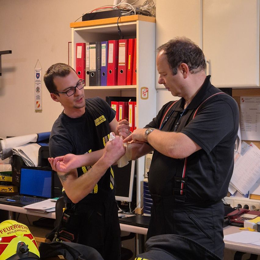 Zwei Männer in Feuerwehruniformen befinden sich in einem Büro. Ein Mann hält den Arm des anderen und wickelt einen Verband um ihn.