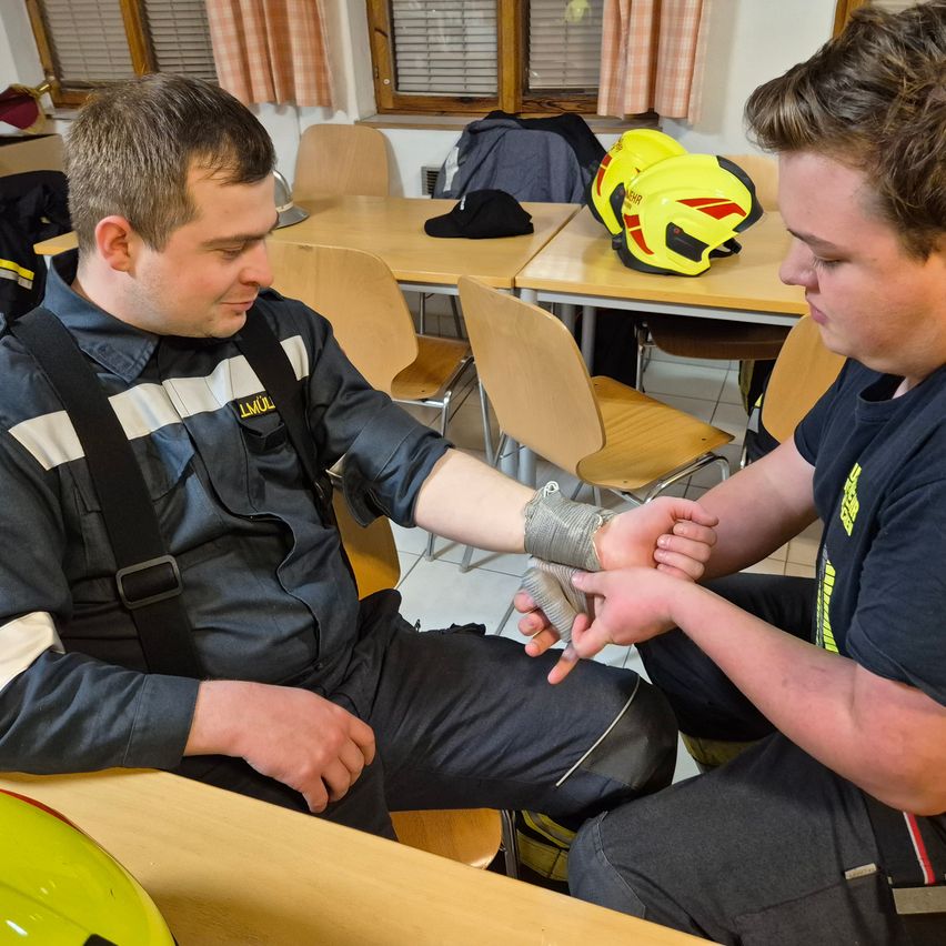 Zwei Männer in Uniformen sind drinnen. Einer hat einen bandagierten Arm und der andere übt Druck aus. In der Nähe stehen Tische mit Helmen und Jacken. Fenster mit Jalousien sind im Hintergrund.