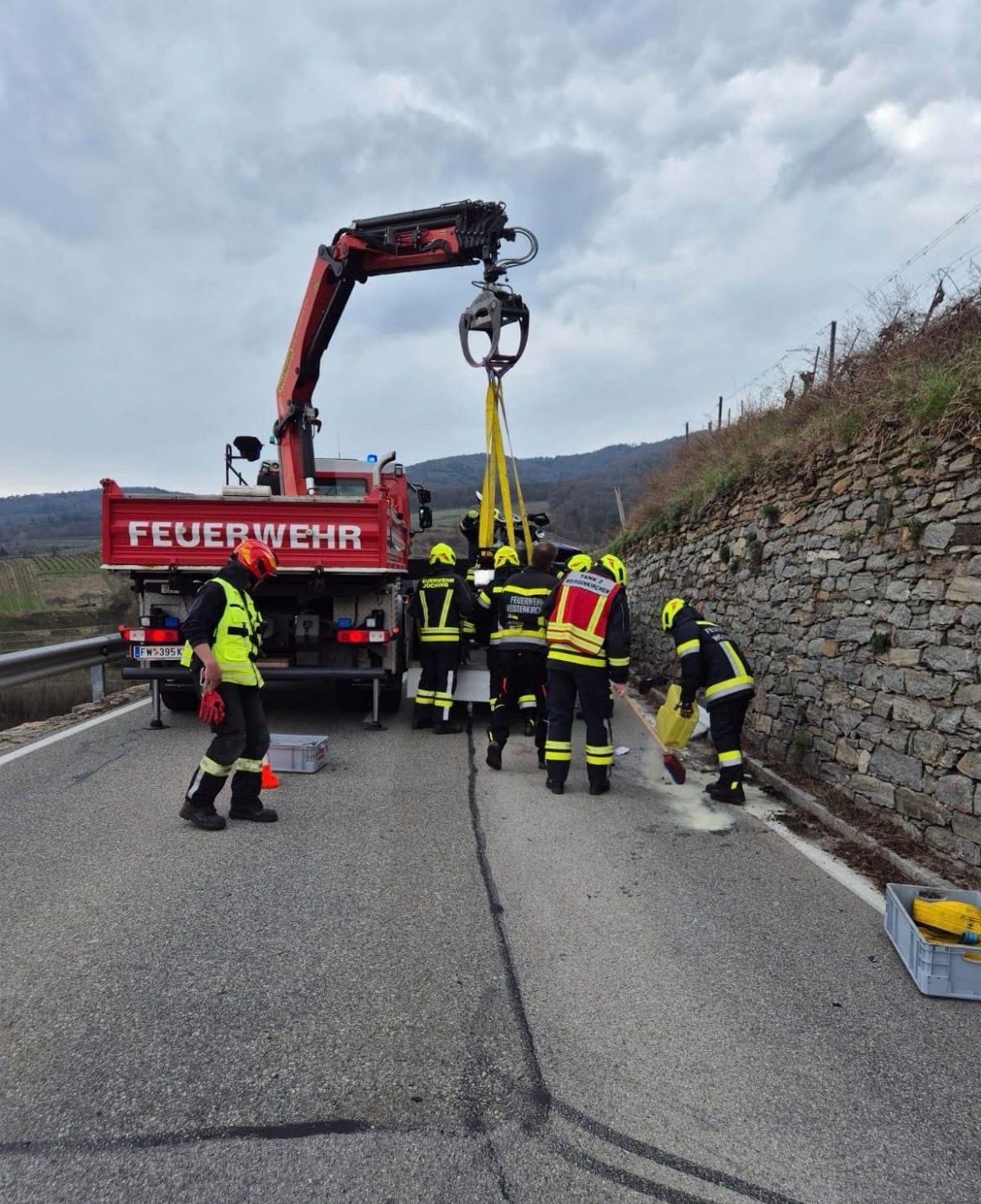 Feuerwehrleute reinigen eine Straße mit einem Kran in der Nähe. Einer hält einen Besen, während ein anderer fegt. Ein Feuerwehrwagen steht mit dem Rücken zur Straße.