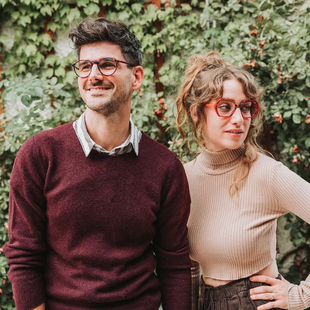 Ein Mann und eine Frau mit Brille stehen lächelnd eng beieinander vor einer Wand mit grünen Blättern und kleinen Früchten.