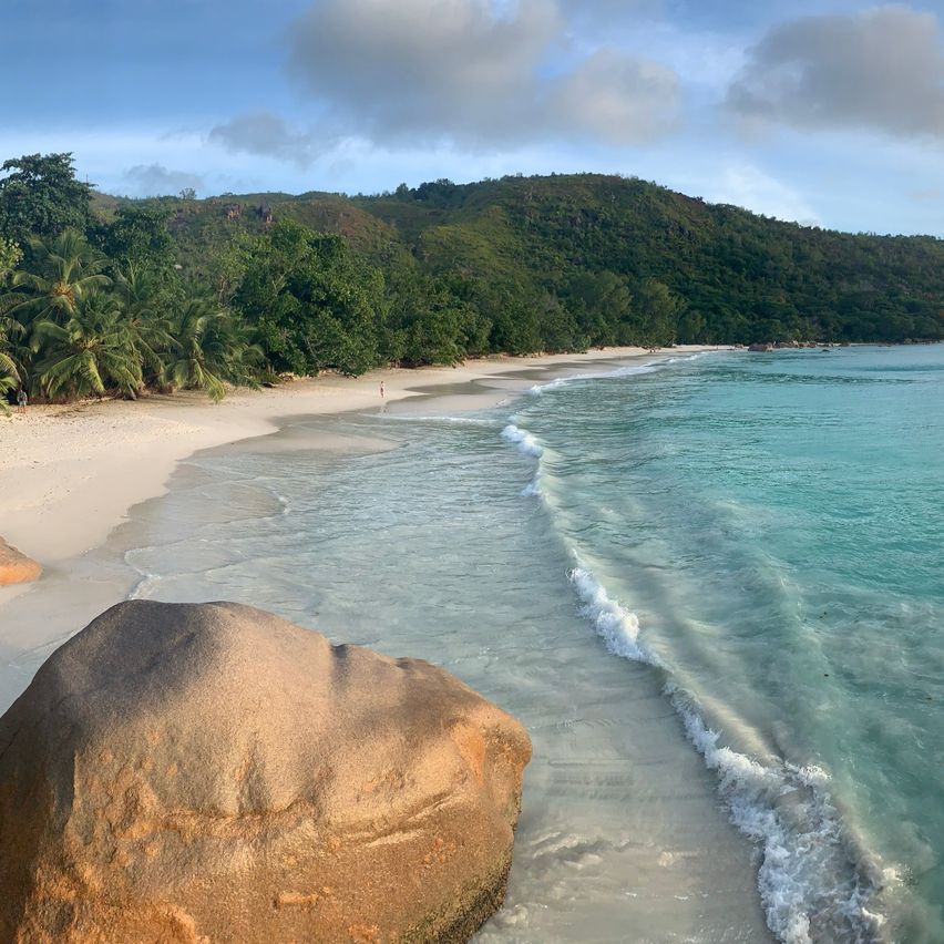 Ein ruhiger Strand mit klarem blauem Wasser und einem großen Felsen am Ufer, umgeben von üppigem grünen Bäumen und einem entfernten Berg unter einem teilweise bewölkten Himmel.