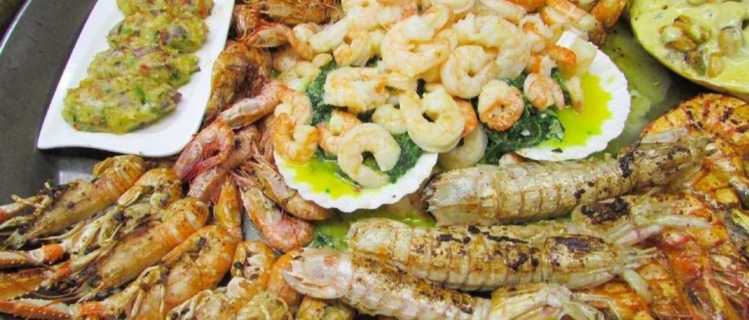 Eine Auswahl an Meeresfrüchten, darunter Garnelen und Hummer, auf weißen Tellern angeordnet. Einige Garnelen sind mit grünem Gemüse serviert.