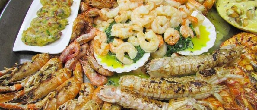 Eine Auswahl an Meeresfrüchten, darunter Garnelen und Hummer, auf weißen Tellern angeordnet. Einige Garnelen sind mit grünem Gemüse serviert.