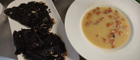 Two white plates with food, one with black food and the other with a creamy soup topped with bacon bits.