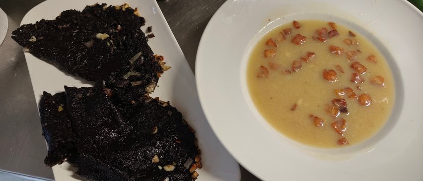Two white plates with food, one with black food and the other with a creamy soup topped with bacon bits.