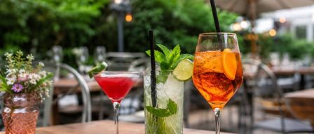 Drei Cocktails auf einem Holztisch in einem Restaurant im Freien, wobei einer eine rote Flüssigkeit, ein anderer ein grünes Blatt und Eis hat und der letzte eine Orangensaft enthält.