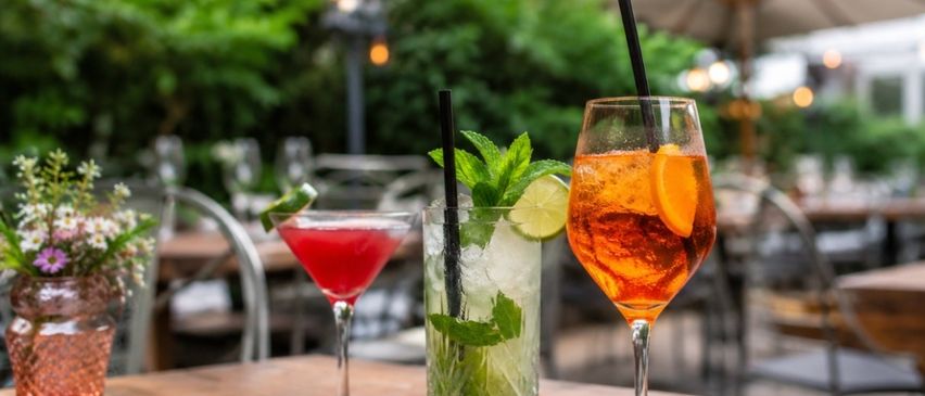 Three cocktails on a wooden table in an outdoor restaurant, with one having a red liquid, another with a green leaf and ice, and the last one with orange juice.