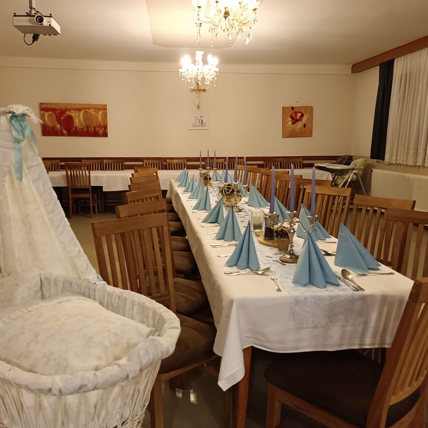 Ein langer Esstisch ist für einen besonderen Anlass gedeckt, geschmückt mit blauen Servietten, Kerzen und Besteck. Eine elegante Kronleuchter hängt von der Decke. Ein Babybett steht in der Nähe.