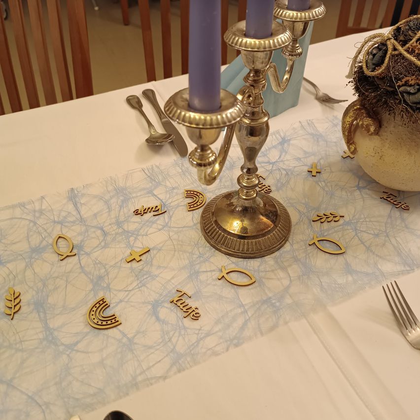 Ein Tisch ist mit einem blau-weißen Tuch gedeckt, das goldene Symbole wie Kreuze und Fische zeigt. Ein Kerzenleuchter mit zwei lila Kerzen steht auf dem Tisch, und ein dekoratives Objekt ist rechts platziert.