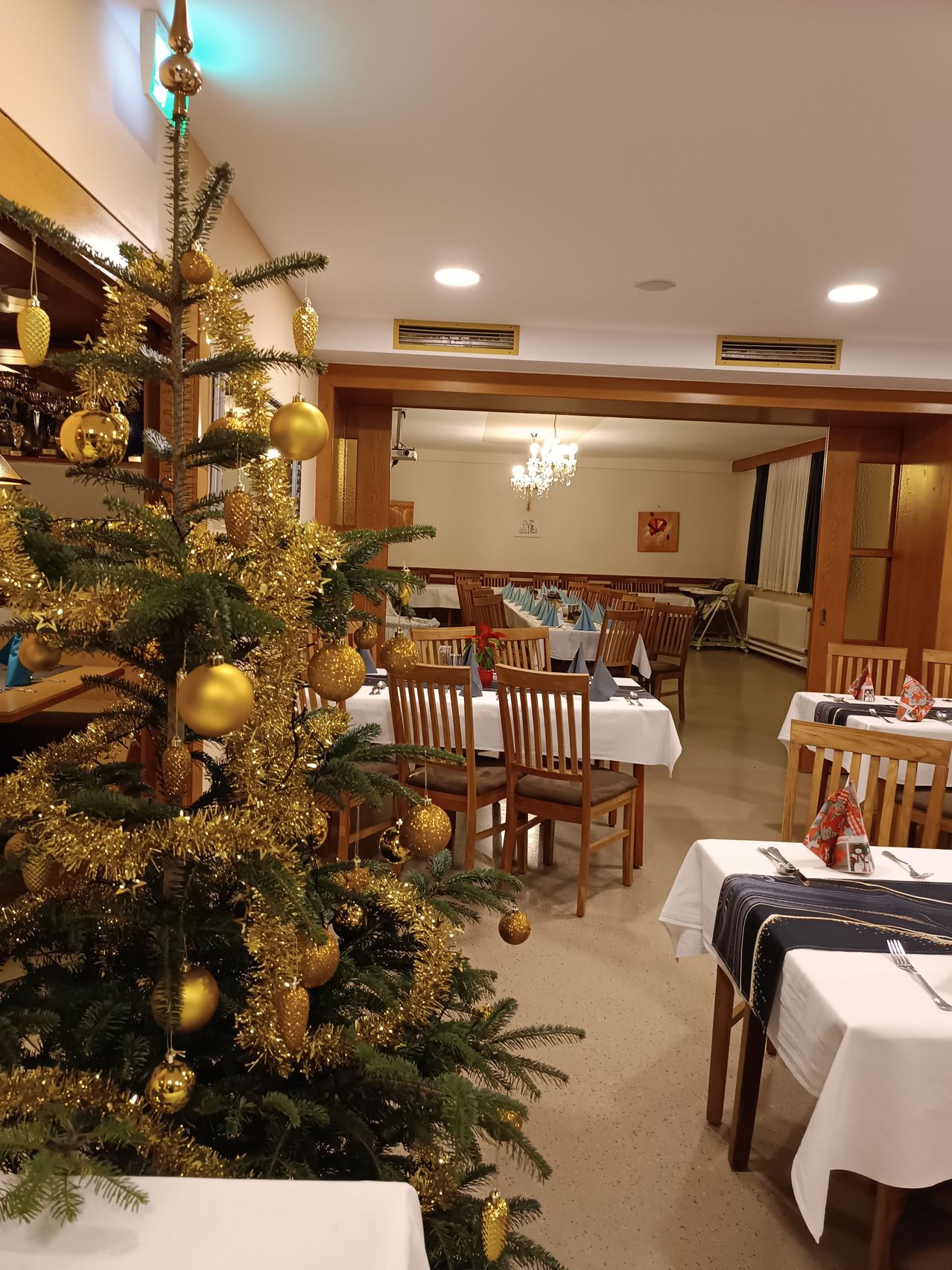 Ein Restaurantinnenraum mit einem geschmückten Weihnachtsbaum, mehreren Tischen mit weißen Tischdecken und Holzstühlen, die für ein festliches Ereignis vorbereitet sind. Der Essbereich ist gut beleuchtet, mit goldenen Ornamenten am Baum und dekorativen Gegenständen auf den Tischen.