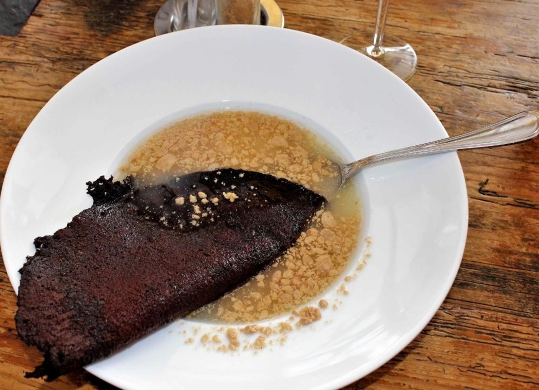 Ein Stück Schokoladenkuchen liegt auf einem weißen Teller, teilweise in einer cremigen Sauce eingetaucht, mit einem Löffel daneben, auf einem Holztisch.