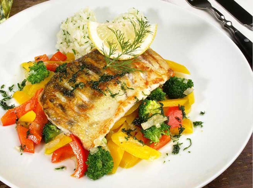 Ein weißer Teller zeigt ein gegrilltes Fischfilet mit Zitrone und Kräutern, begleitet von einem Bett aus buntem Gemüse wie Brokkoli, Paprika und Zwiebeln, mit Reis als Beilage.