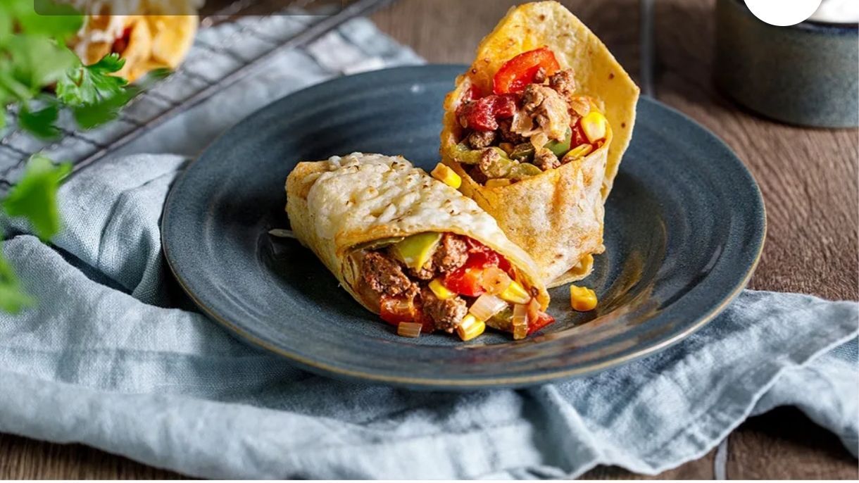 Ein Teller mit zwei Burritos, jeder mit einer Mischung aus Hackfleisch, Tomaten, Paprika, Zwiebeln und Mais, eingewickelt in goldene Tortillas, auf einem blauen Teller mit einem blauen Tuch im Hintergrund.