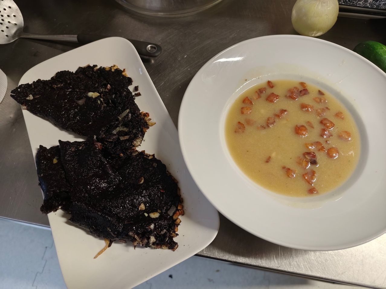 Zwei weiße Teller mit schwarzem Essen auf einem, eine cremige Suppe mit Speckstückchen auf dem anderen und einer Zwiebel auf dem Tisch.