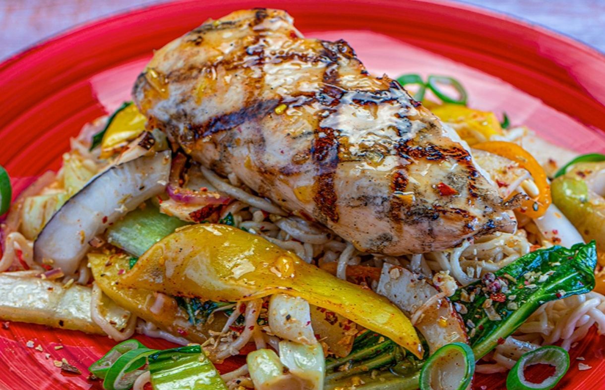 Grilled chicken breast atop a bed of noodles, accompanied by grilled vegetables, resting on a red plate.