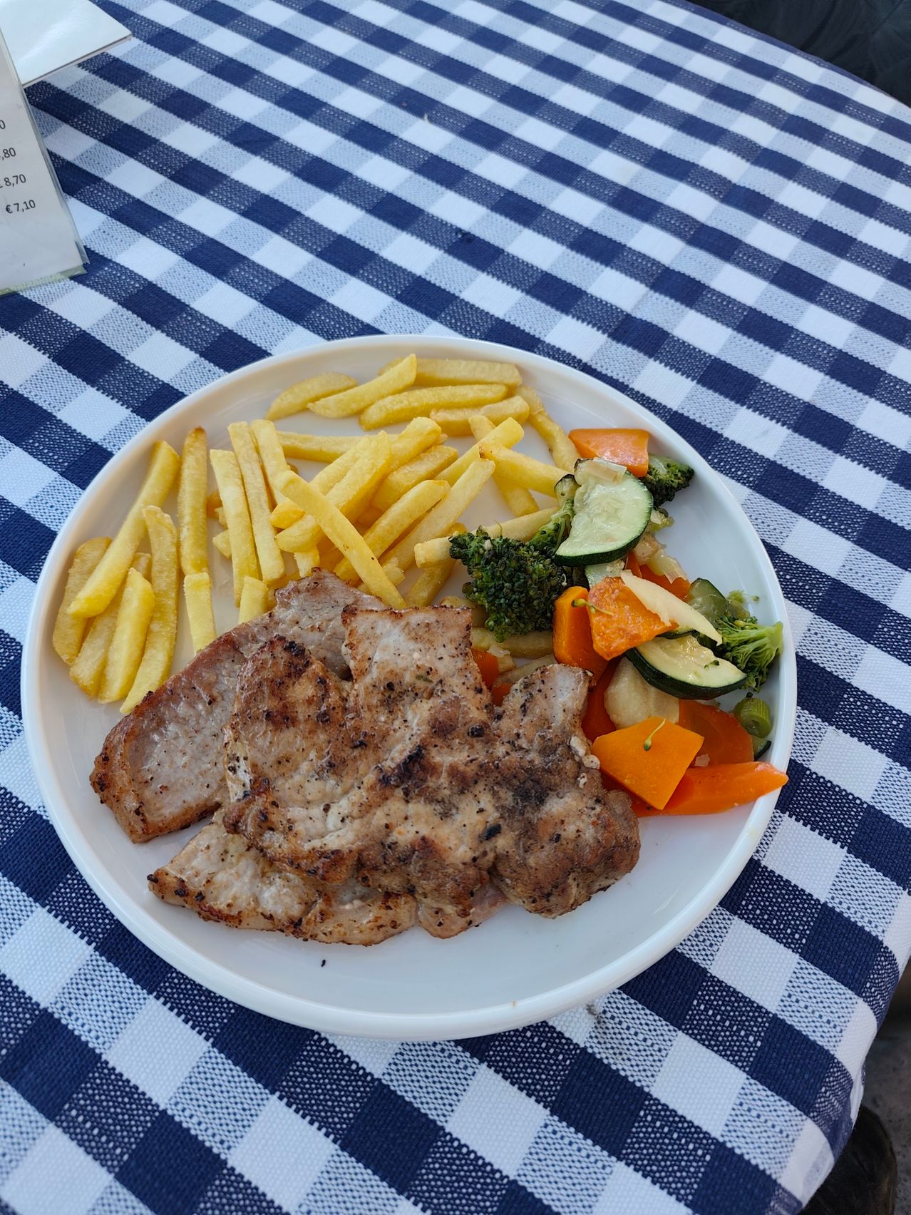 Ein Teller mit gegrillten Schweinekoteletts, Pommes Frites und einer Mischung aus gedämpften Gemüsen auf einer blau-weiß karierten Tischdecke.