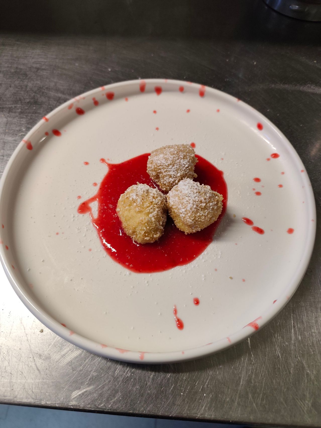 A white plate with three golden brown balls on red sauce. The plate has a white border and red splashes. The dessert is on a metallic surface.