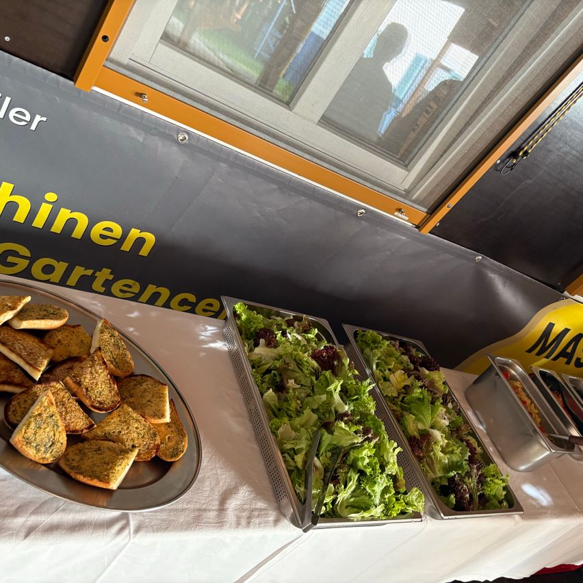 Ein Tisch zeigt Essen: ein Teller mit Knoblauchbrot, zwei Tabletts mit Salat und zwei Tabletts mit anderen Gerichten. Die Wand dahinter hat ein Banner mit der Aufschrift 'Garten' und ein Fenster mit Blick auf eine Person.
