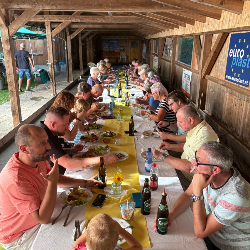 Eine große Gruppe von Menschen sitzt an einem langen Tisch und genießt ihre Mahlzeiten. Der Tisch ist mit einer gelben Tischdecke bedeckt, und Flaschen und Gläser sind darauf angeordnet. Ein Schild mit dem Text 'euro plast' ist an der Wand hinter ihnen montiert. Eine Person geht auf der linken Seite.