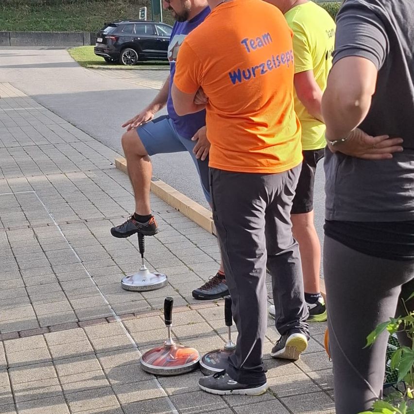 Eine Gruppe von Männern steht auf einer Ziegelsteinpflasterung, einer balanciert auf einem Einrad. Der Mann im orangefarbenen Shirt sagt 'Team Wurzelsepp'. Ein schwarzer SUV ist am Straßenrand geparkt.