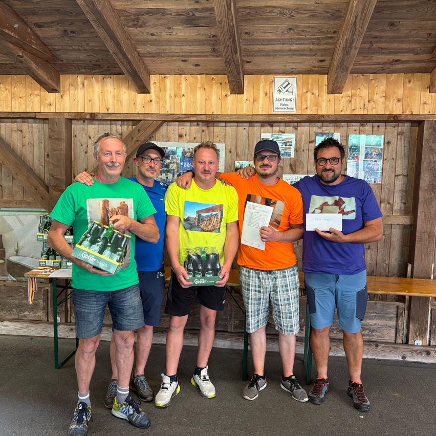 Fünf Männer stehen in einem Raum mit Holzwänden, lächeln und posieren für ein Foto. Sie halten Gegenstände und Urkunden. Ein Holztisch und eine Bank sind im Hintergrund.