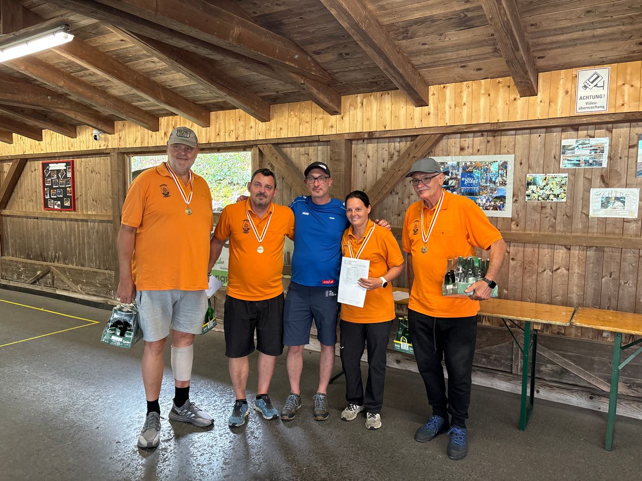 Fünf Personen mit orangefarbenen Shirts und Medaillen stehen in einem Raum mit Holzwänden und einem Tisch mit Flaschen.