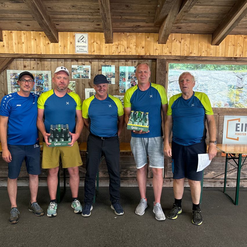 Fünf Männer stehen in einem Raum mit Holzwänden und lächeln für ein Foto. Sie halten Kisten mit Bier. Zwei Männer tragen Brillen und zwei tragen Hüte. Ein Fenster zeigt einen großen Felsen.