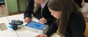 Zwei junge Mädchen sitzen an einem Tisch, eine konzentriert sich auf ein Tablet, während die andere es berührt. Auf dem Tisch steht ein kleiner Roboter.