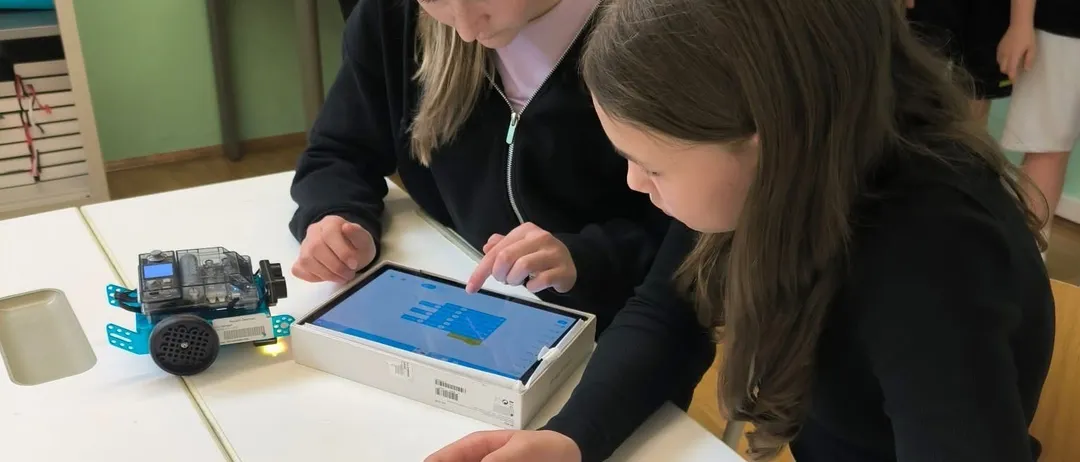 Zwei junge Mädchen sitzen an einem Tisch, eine konzentriert sich auf ein Tablet, während die andere es berührt. Auf dem Tisch steht ein kleiner Roboter.