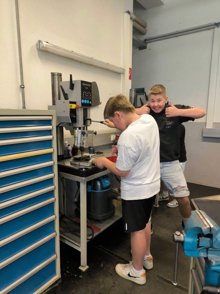 Zwei Jungen arbeiten in einer Werkstatt an einer Maschine. Einer bedient die Maschine, während der andere Daumen hochhält.