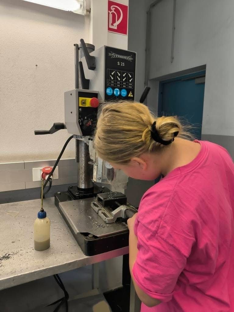 Eine Frau in einem rosa T-Shirt arbeitet in einer Werkstatt an einer Maschine. Sie beugt sich über einen Arbeitstisch mit einer Flasche Flüssigkeit und einem Werkzeug in der Hand.