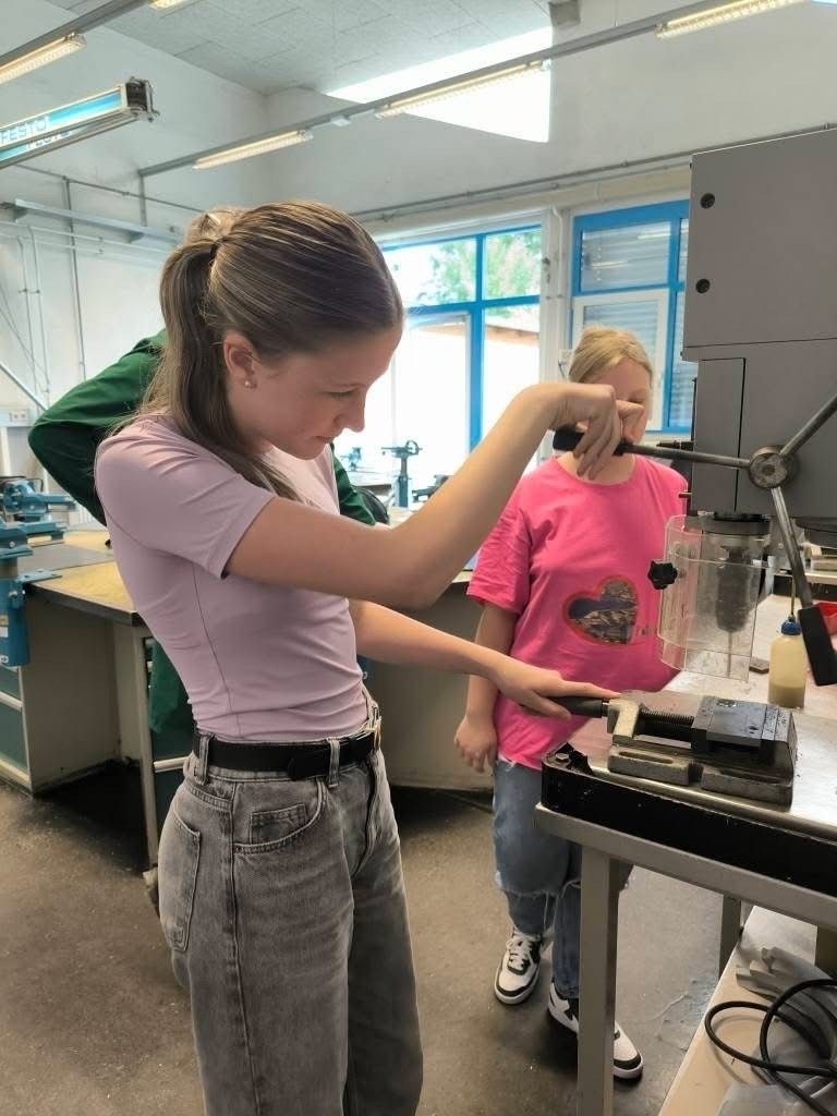 Ein Mädchen in einem rosa T-Shirt bohrt mit einer Bohrmaschine ein Loch in ein Metallstück, während ein anderes Mädchen in einem rosa T-Shirt zusieht.