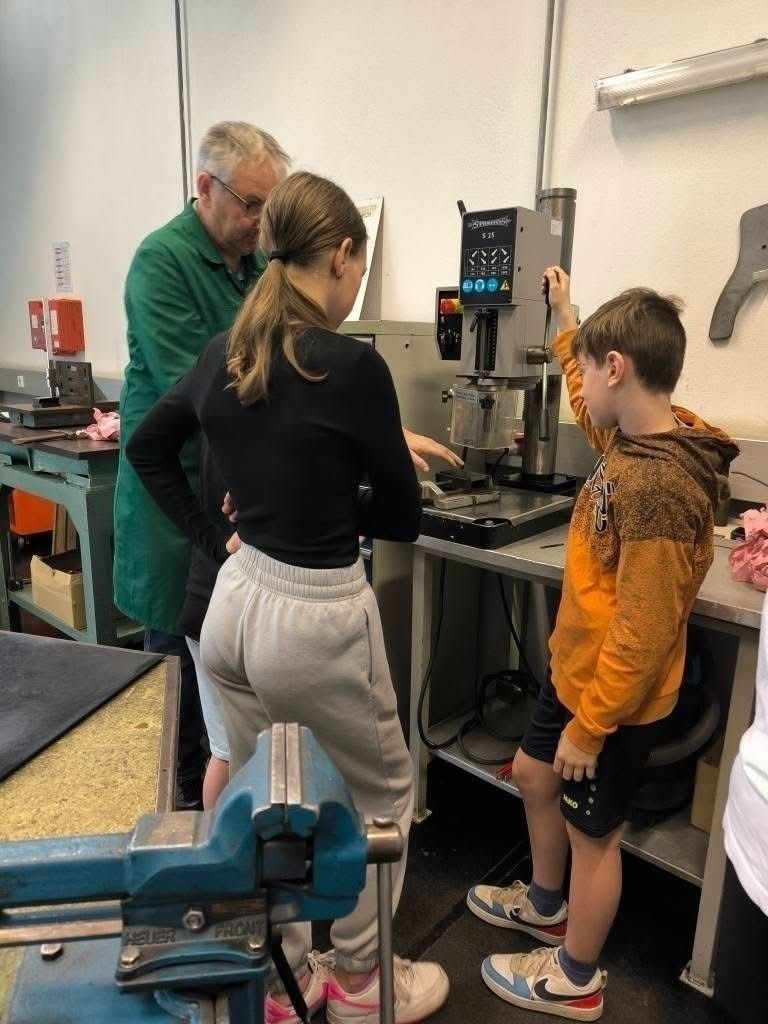Drei Personen befinden sich in einer Werkstatt. Eine Frau bedient eine Maschine, während ein Mann und ein Junge zusehen.