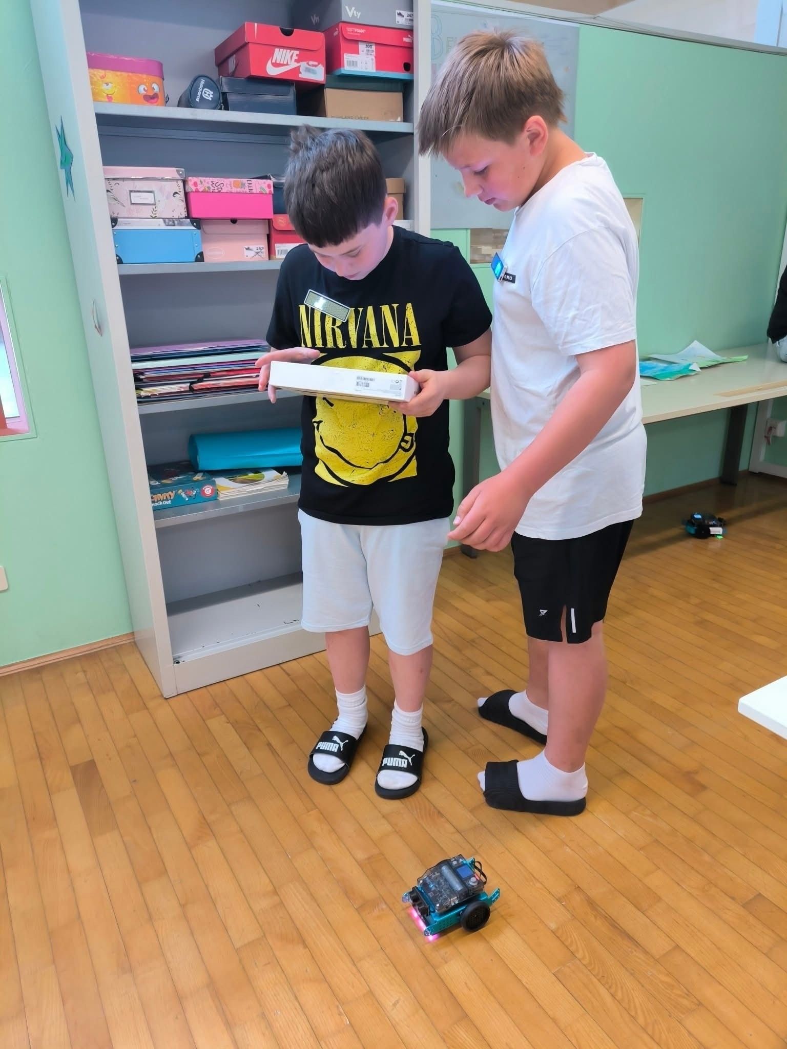 Zwei Jungen stehen in einem Klassenzimmer und betrachten eine weiße Box. Einer von ihnen trägt ein schwarzes T-Shirt mit einem gelben Design. Hinter ihnen steht ein Regal mit vielen Gegenständen. Auf der rechten Seite befindet sich ein Tisch mit Büchern und Papieren.