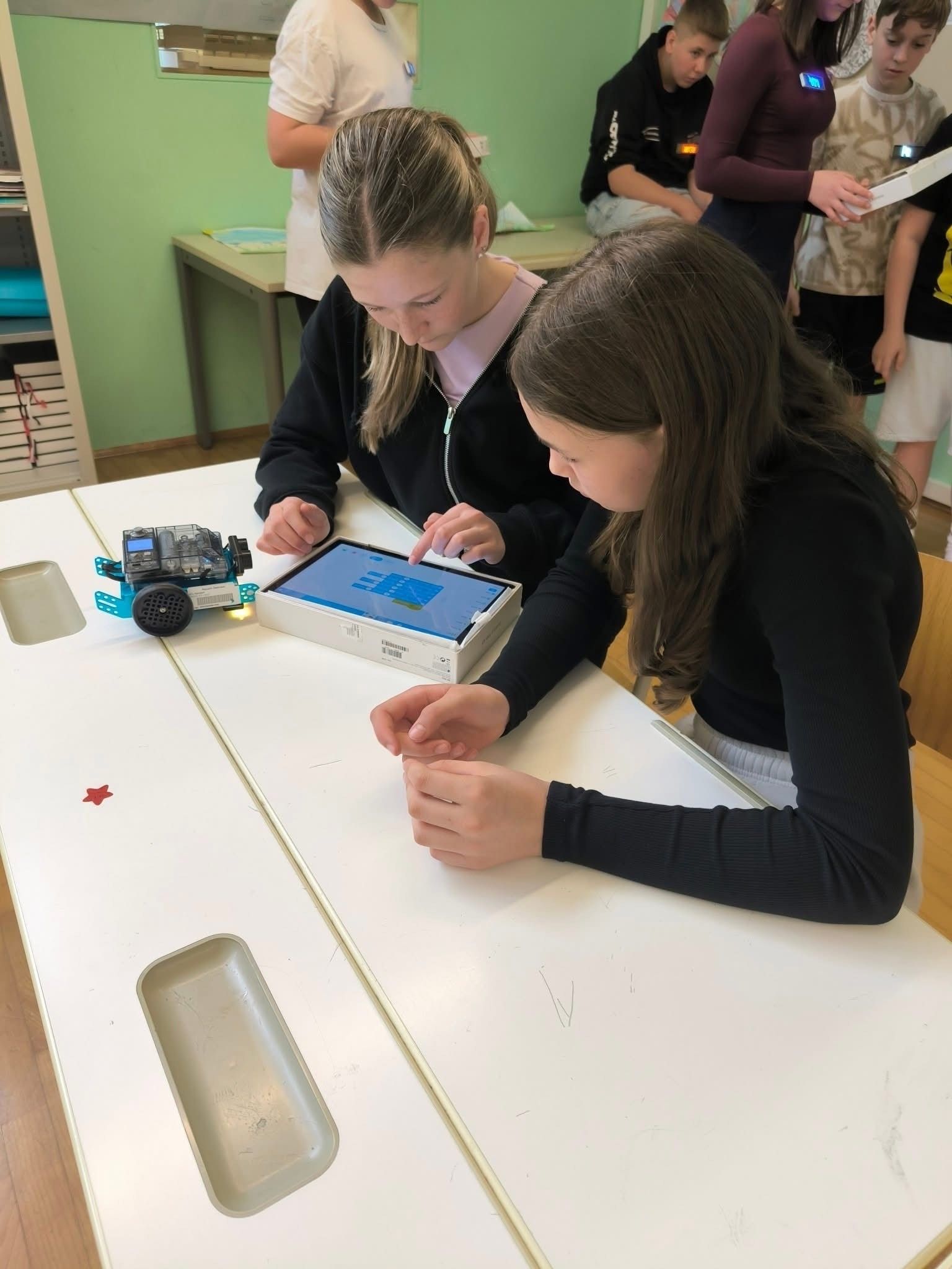 Zwei Mädchen sitzen an einem Tisch im Klassenzimmer, eines der Mädchen tippt auf einem Tablet, während das andere Mädchen auf den Bildschirm schaut. Ein kleiner Roboter sitzt auf dem Tisch.