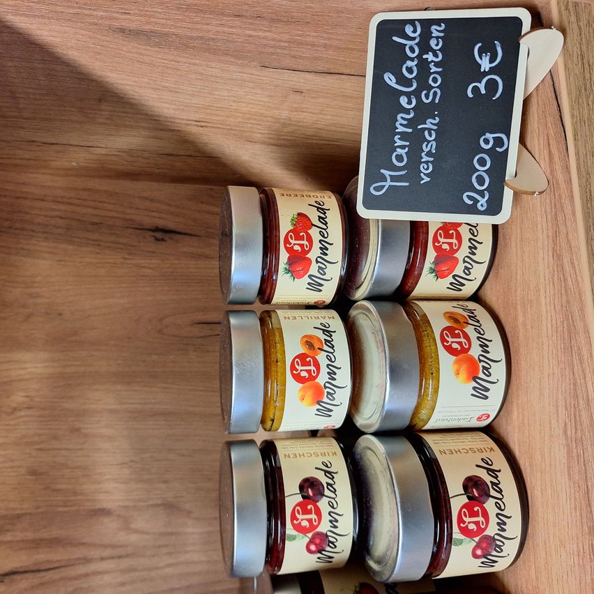 Mehrere Glasgläser mit Erdbeer-, Aprikosen- und Kirschmarmelade auf einem Holzregal. Ein Tafel mit dem Text 'Verschee Sorten' und '3,60€' ist ebenfalls zu sehen.