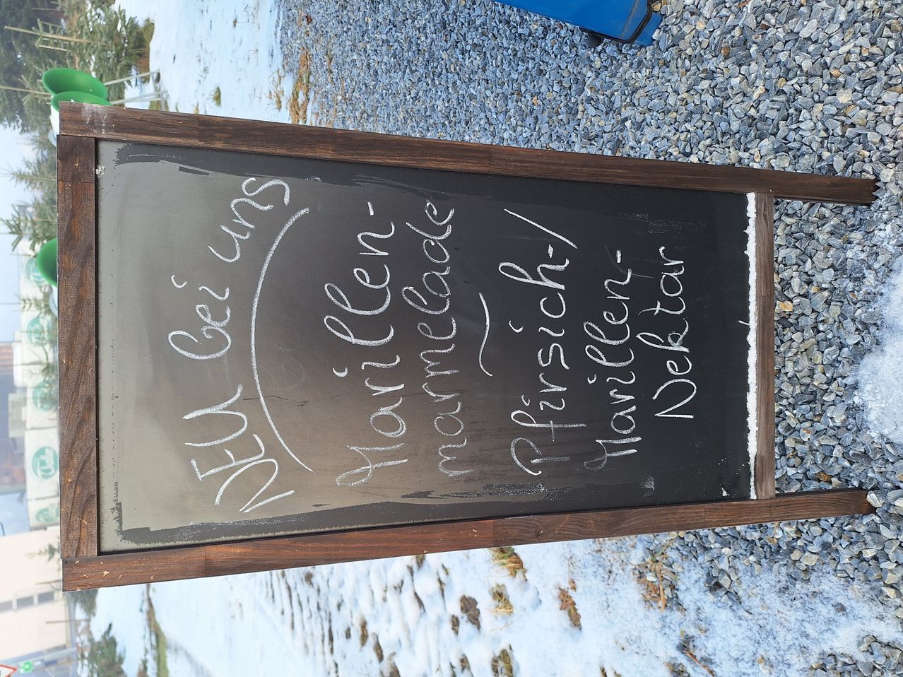 Ein Kreidetafel mit handgeschriebenem Text steht auf einer felsigen Oberfläche. Der Text enthält Namen von Gerichten wie 'Morilles', 'Peters Fisch', 'Hummerchen' und 'Delkat'. Der Hintergrund zeigt eine verschneite Landschaft.